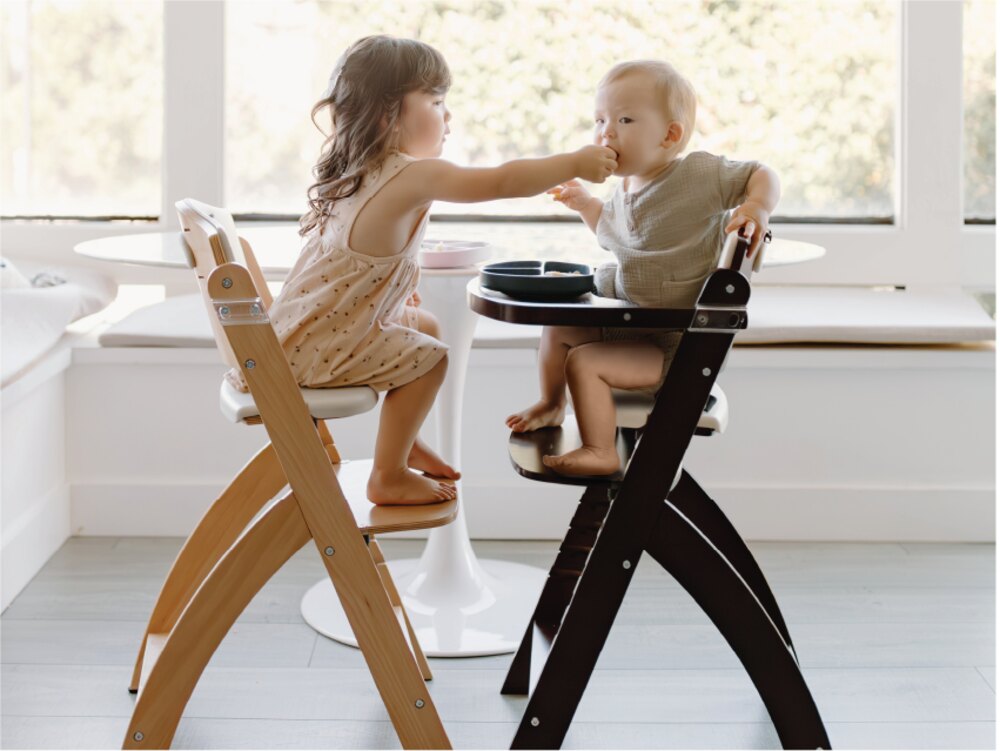 Sister feeding baby brother in wooden high chairs