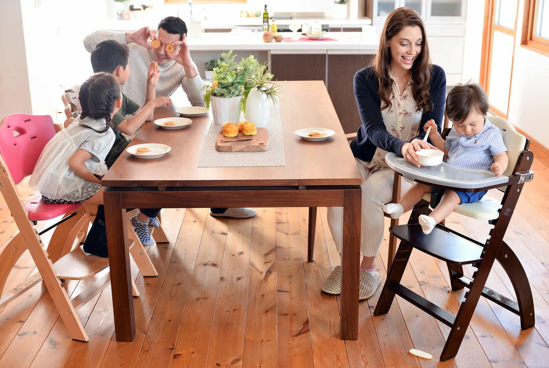 Family at a dining table with two children sitting in adjustable wooden high chairs and a toddler in a high chair on the right while a parent feeds the toddler.