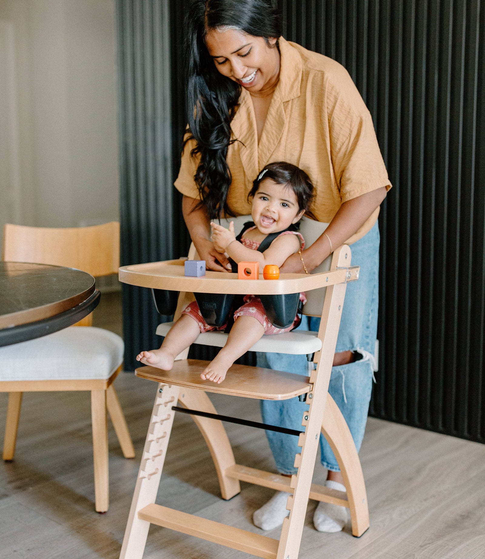 Smiling baby seated in a wooden Abiie high chair with a tray holding colorful blocks, with a parent standing behind adjusting the child’s seat.
